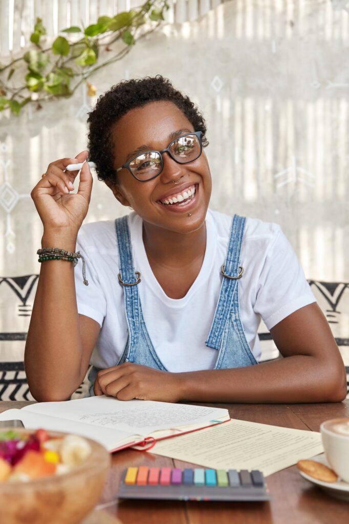 Photo of cheerful black woman with broad smile, ponders on idea for solution, plans costs and writes