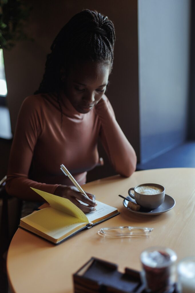 Portrait of young African-American woman sitting at table with cup of coffee in cafe, writing text